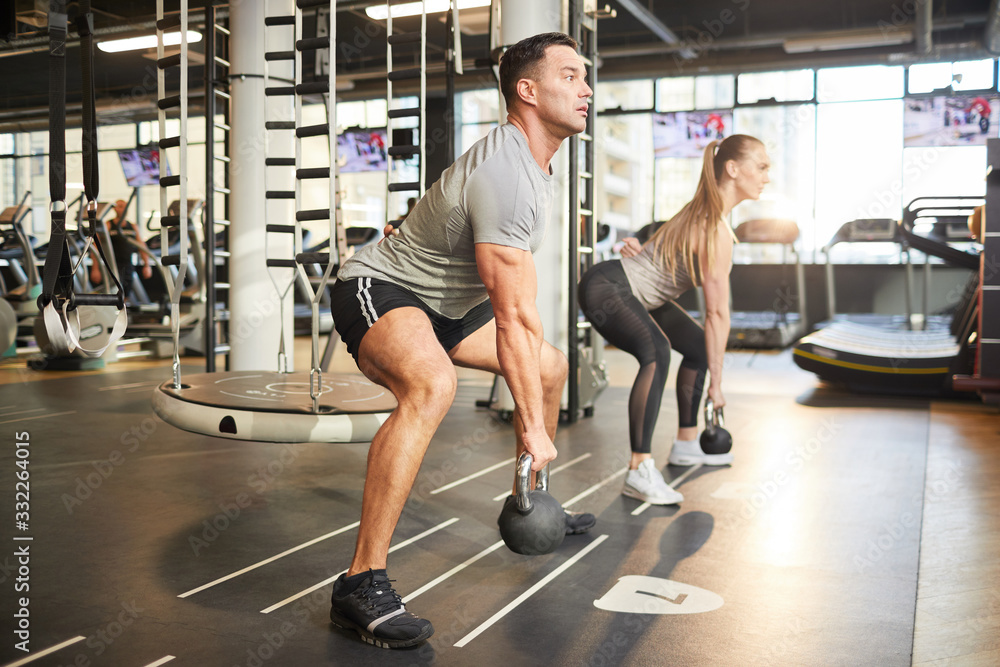 Side view portrait of muscular couple swinging kettlebells during strength workout in modern gym, copy space