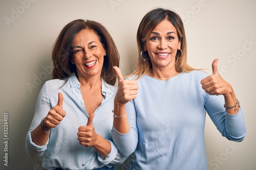 Middle age beautiful couple of sisters standing over isolated white background success sign doing positive gesture with hand, thumbs up smiling and happy. Cheerful expression and winner gesture.