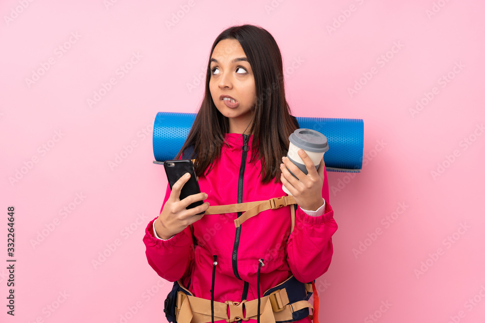 Young mountaineer girl with a big backpack over isolated pink background holding coffee to take away and a mobile while thinking something