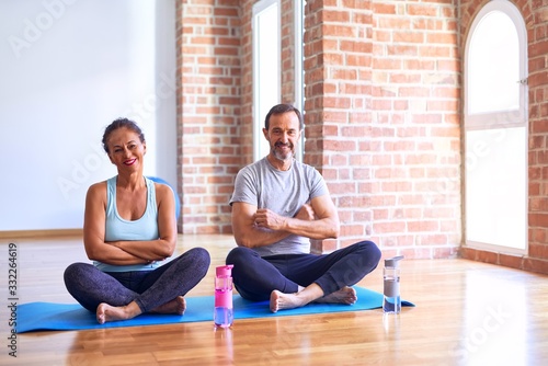 Middle age sporty couple sitting on mat doing stretching yoga exercise at gym happy face smiling with crossed arms looking at the camera. Positive person.