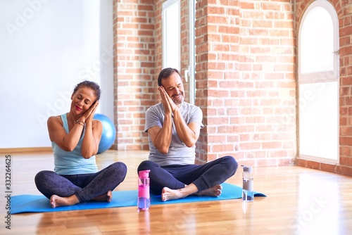 Middle age sporty couple sitting on mat doing stretching yoga exercise at gym sleeping tired dreaming and posing with hands together while smiling with closed eyes.