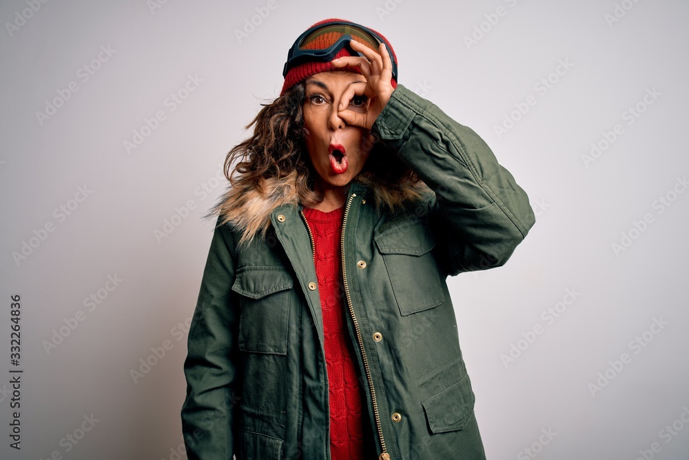 Middle age skier woman wearing snow sportswear and ski goggles over white background doing ok gesture shocked with surprised face, eye looking through fingers. Unbelieving expression. Middle age skier woman wearing snow sportswear and ski goggles over white background doing ok gesture shocked with surprised face, eye looking through fingers. Unbelieving expression.