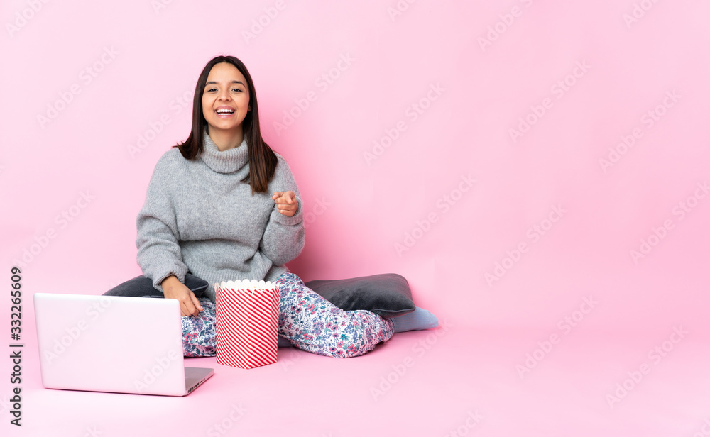Young mixed race woman eating popcorn while watching a movie on the laptop surprised and pointing front Young mixed race woman eating popcorn while watching a movie on the laptop surprised and pointing front
