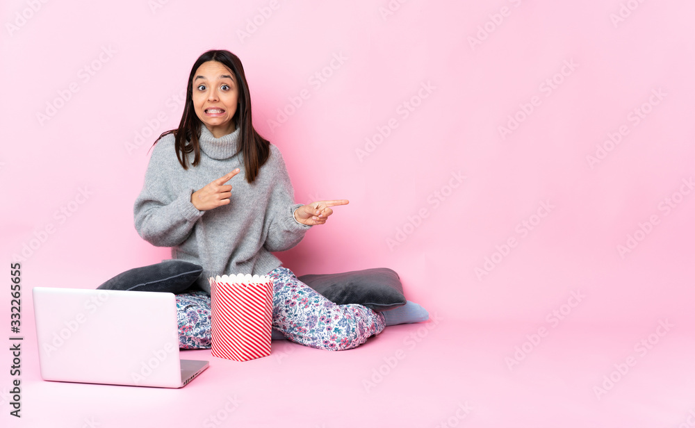 Young mixed race woman eating popcorn while watching a movie on the laptop frightened and pointing to the side Young mixed race woman eating popcorn while watching a movie on the laptop frightened and pointing to the side