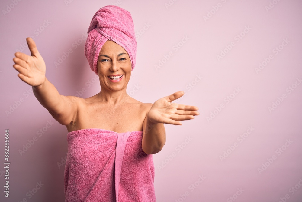 Middle age woman wearing bath towel from beauty body care over pink background looking at the camera smiling with open arms for hug. Cheerful expression embracing happiness. Middle age woman wearing bath towel from beauty body care over pink background looking at the camera smiling with open arms for hug. Cheerful expression embracing happiness.