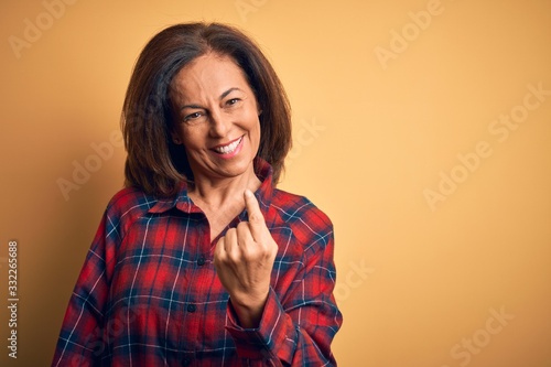 Middle age beautiful woman wearing casual shirt standing over isolated yellow background Beckoning come here gesture with hand inviting welcoming happy and smiling