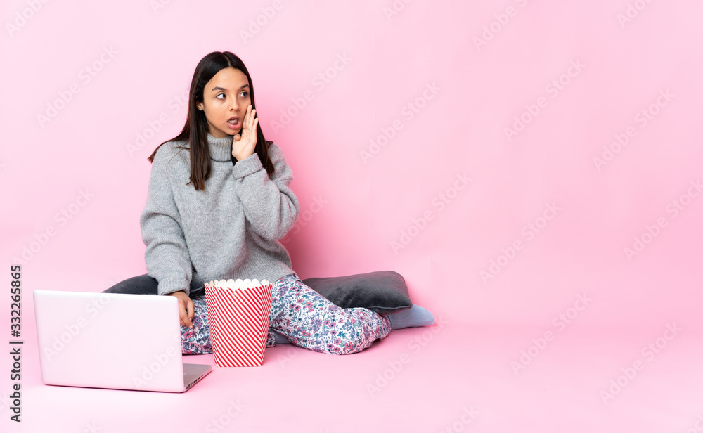 Young mixed race woman eating popcorn while watching a movie on the laptop whispering something with surprise gesture while looking to the side Young mixed race woman eating popcorn while watching a movie on the laptop whispering something with surprise gesture while looking to the side