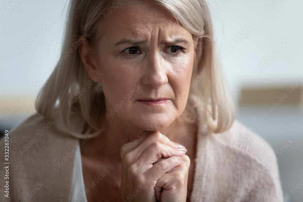 Frowning upset mature woman put head on folded hands, looking in distance. Lost in thoughts upset woman sitting alone, head shot close up portrait. Mental disorder or dementia, health problem concept. Frowning upset mature woman put head on folded hands, looking in distance. Lost in thoughts upset woman sitting alone, head shot close up portrait. Mental disorder or dementia, health problem concept.