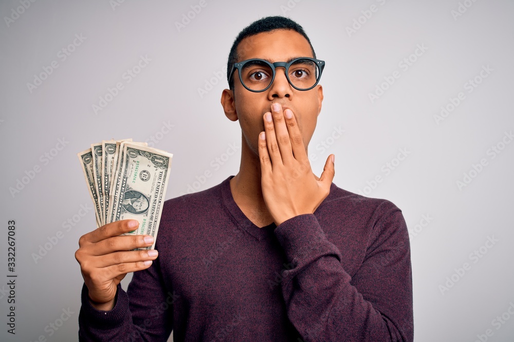 Handsome african american man holding bunch of dollars banknotes over white background cover mouth with hand shocked with shame for mistake, expression of fear, scared in silence, secret concept Handsome african american man holding bunch of dollars banknotes over white background cover mouth with hand shocked with shame for mistake, expression of fear, scared in silence, secret concept