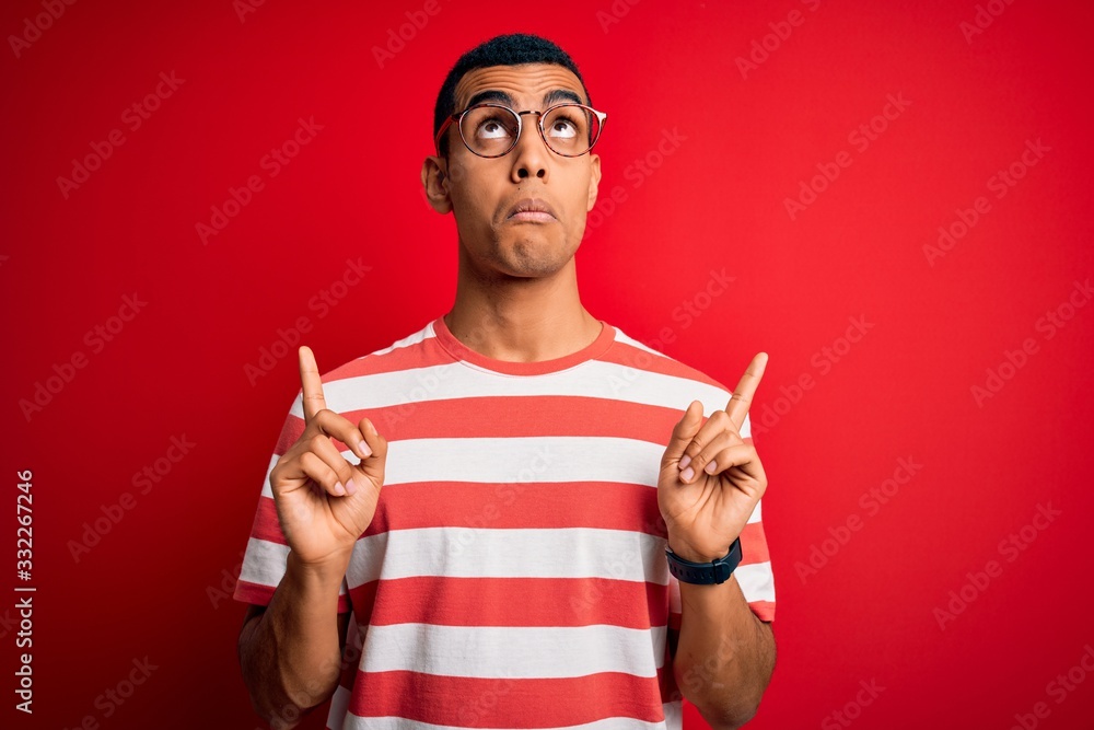Fototapeta premium Young handsome african american man wearing casual striped t-shirt and glasses Pointing up looking sad and upset, indicating direction with fingers, unhappy and depressed.