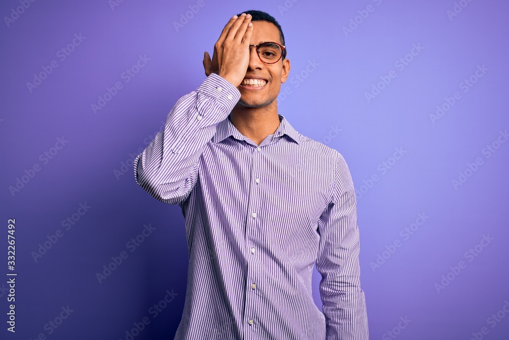 Handsome african american man wearing striped shirt and glasses over purple background covering one eye with hand, confident smile on face and surprise emotion. Handsome african american man wearing striped shirt and glasses over purple background covering one eye with hand, confident smile on face and surprise emotion.
