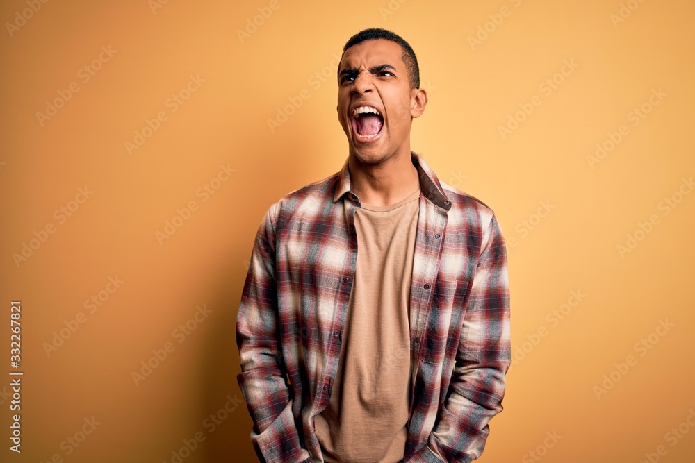 Young handsome african american man wearing casual shirt standing over yellow background angry and mad screaming frustrated and furious, shouting with anger. Rage and aggressive concept. Young handsome african american man wearing casual shirt standing over yellow background angry and mad screaming frustrated and furious, shouting with anger. Rage and aggressive concept.