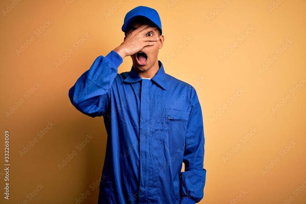Young african american mechanic man wearing blue uniform and cap over yellow background peeking in shock covering face and eyes with hand, looking through fingers with embarrassed expression.