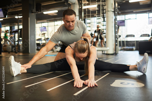 Full length portrait of muscular coach helping young woman doing splits and stretching exercises during fitness workout in gym, copy space