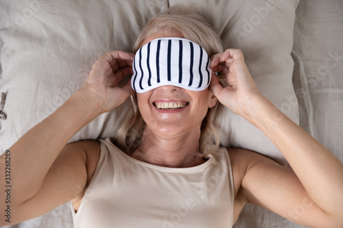 Above top view smiling middle aged woman wearing sleeping mask, feeling energetic after good night rest. Head shot close up satisfied pleased relaxed older granny enjoying weekend morning in bed.