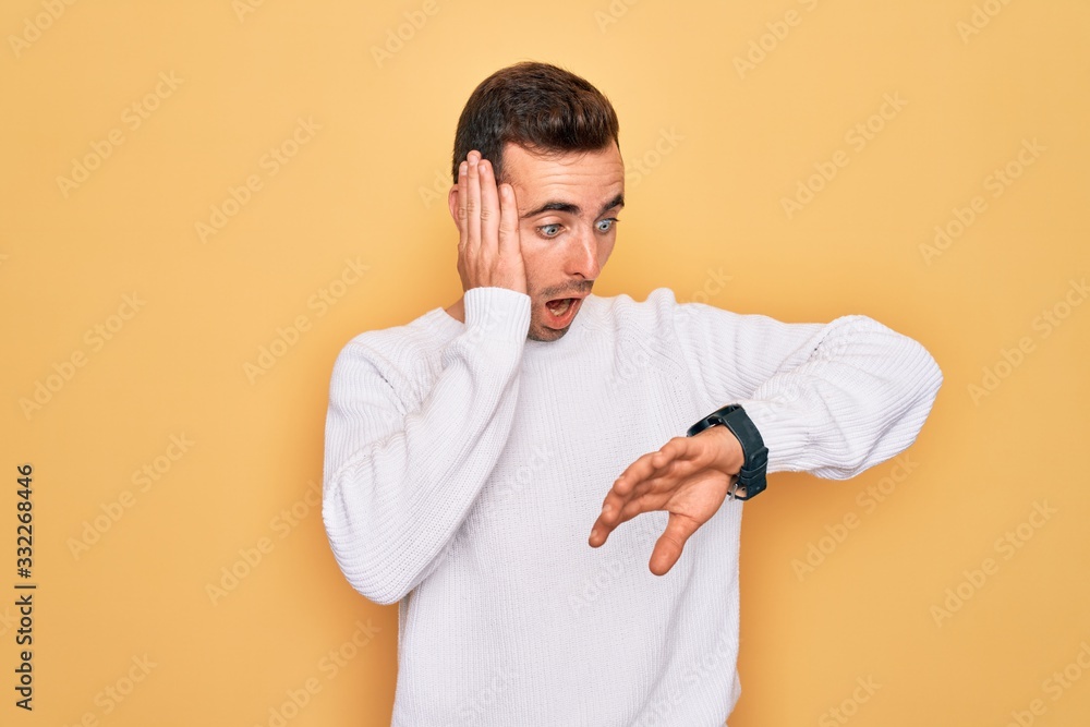 Young handsome man with blue eyes wearing casual sweater standing over yellow background Looking at the watch time worried, afraid of getting late Young handsome man with blue eyes wearing casual sweater standing over yellow background Looking at the watch time worried, afraid of getting late