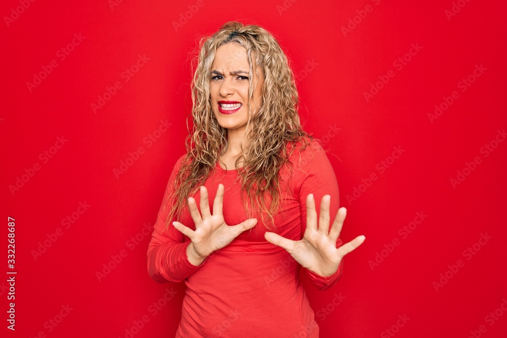Young beautiful blonde woman wearing red casual t-shirt standing over isolated background disgusted expression, displeased and fearful doing disgust face because aversion reaction. With hands raised