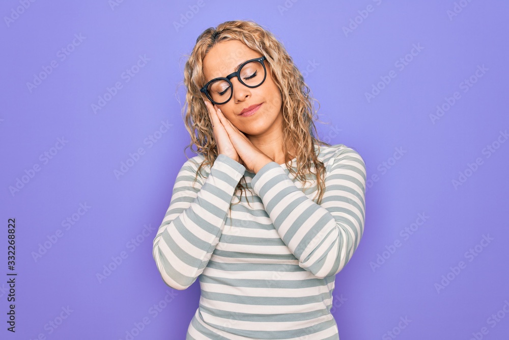 Beautiful blonde woman wearing casual striped t-shirt and glasses over purple background sleeping tired dreaming and posing with hands together while smiling with closed eyes.