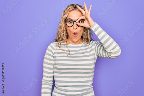 Beautiful blonde woman wearing casual striped t-shirt and glasses over purple background doing ok gesture shocked with surprised face  eye looking through fingers. Unbelieving expression.