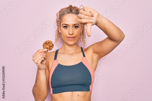 Young beautiful blonde sporty woman doing sport holding cookie over isolated pink background with angry face, negative sign showing dislike with thumbs down, rejection concept