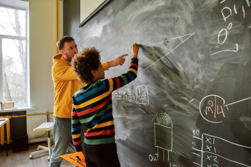 The great aim of education is not knowledge but action. Young male science teacher explaining main rules and formulas in physics and mechanics. A boy is drawing on the blackboard