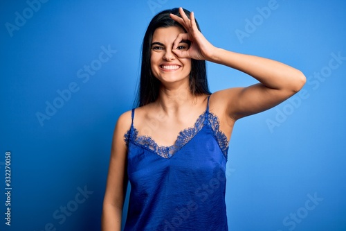 Young beautiful brunette woman wearing elegant and comfortable blue lingerie underwear doing ok gesture with hand smiling, eye looking through fingers with happy face.