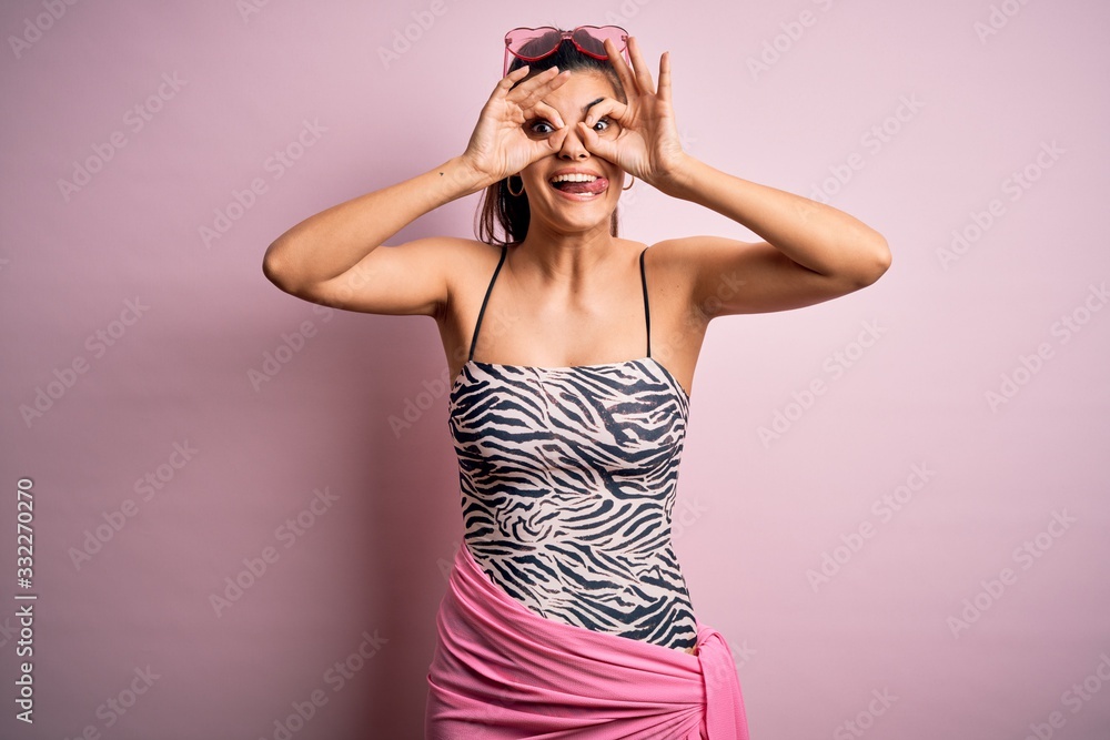 Young beautiful brunette woman on vacation wearing swimsuit over pink background doing ok gesture like binoculars sticking tongue out, eyes looking through fingers. Crazy expression. Young beautiful brunette woman on vacation wearing swimsuit over pink background doing ok gesture like binoculars sticking tongue out, eyes looking through fingers. Crazy expression.