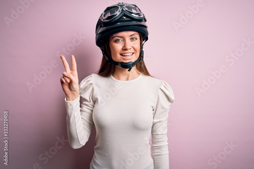 Young beautiful motorcyclist woman with blue eyes wearing moto helmet over pink background smiling looking to the camera showing fingers doing victory sign. Number two.