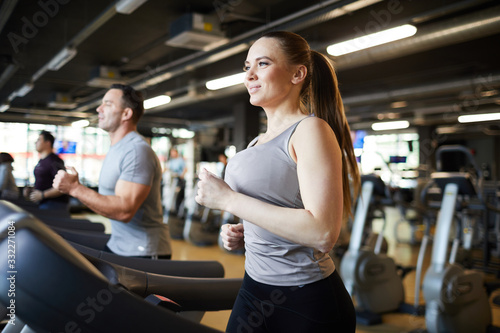 Waist up portrait of smiling young woman running on treadmill while enjoying cardio workout in modern gym, copy space