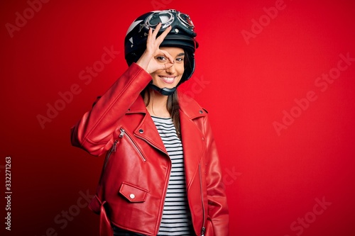 Young beautiful brunette motorcyclist woman wearing motorcycle helmet and red jacket doing ok gesture with hand smiling, eye looking through fingers with happy face.