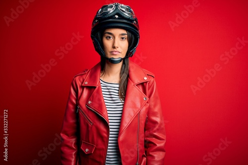 Young beautiful brunette motorcyclist woman wearing motorcycle helmet and red jacket with serious expression on face. Simple and natural looking at the camera. © Krakenimages.com