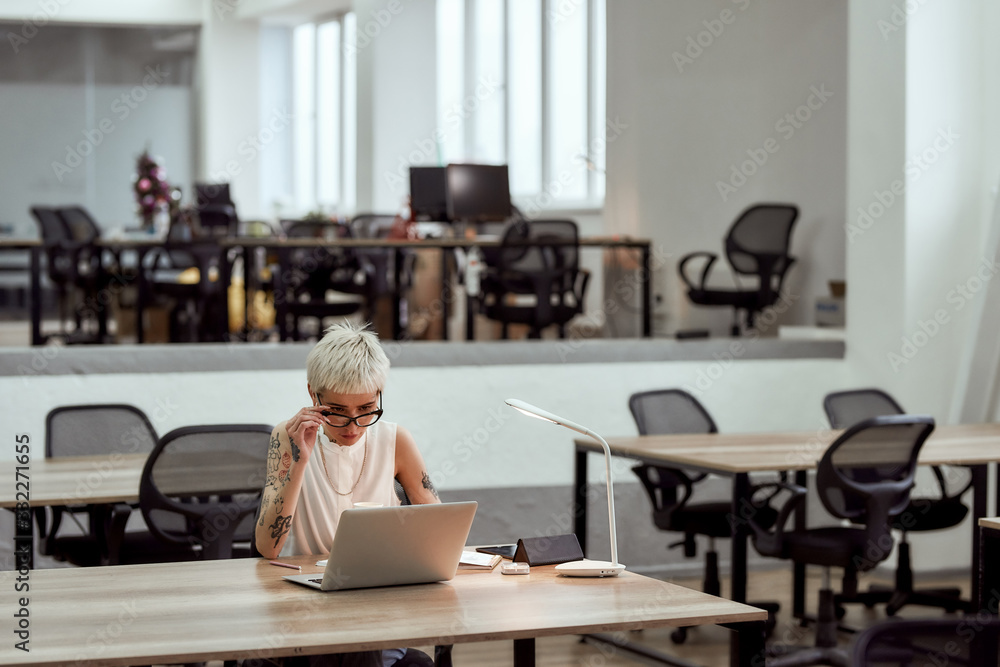 Business lady at work. Young focused blonde tattooed woman in eyewear working with laptop while sitting alone in the modern office Business lady at work. Young focused blonde tattooed woman in eyewear working with laptop while sitting alone in the modern office