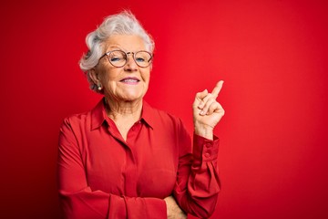 Senior beautiful grey-haired woman wearing casual shirt and glasses over red background with a big smile on face, pointing with hand and finger to the side looking at the camera.