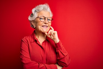 Senior beautiful grey-haired woman wearing casual shirt and glasses over red background looking confident at the camera with smile with crossed arms and hand raised on chin. Thinking positive.