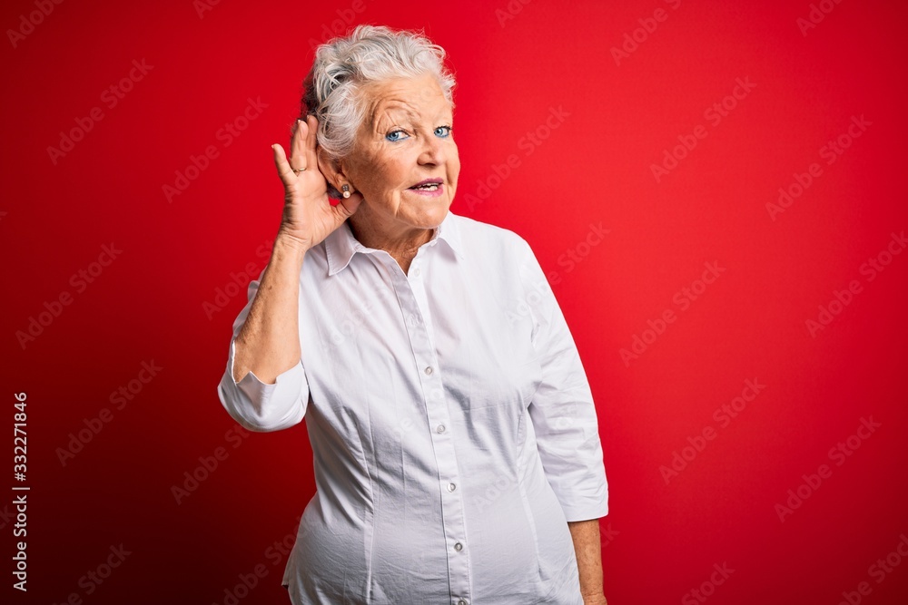 Senior beautiful woman wearing elegant shirt standing over isolated red background smiling with hand over ear listening an hearing to rumor or gossip. Deafness concept. Senior beautiful woman wearing elegant shirt standing over isolated red background smiling with hand over ear listening an hearing to rumor or gossip. Deafness concept.