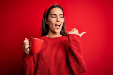 Young beautiful brunette woman drinking mug of coffe over isolated red background pointing and showing with thumb up to the side with happy face smiling