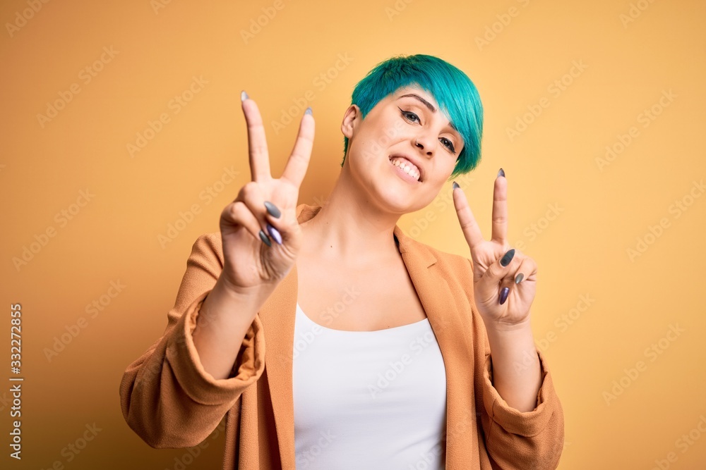 Obraz premium Young beautiful woman with blue fashion hair wearing casual jacket over yellow background smiling looking to the camera showing fingers doing victory sign. Number two.