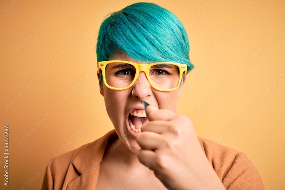 Young beautiful woman with blue fashion hair wearing casual glasses over yellow background annoyed and frustrated shouting with anger, crazy and yelling with raised hand, anger concept