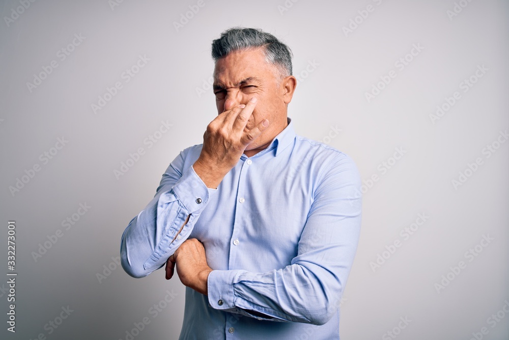 Middle age handsome grey-haired business man wearing elegant shirt over white background smelling something stinky and disgusting, intolerable smell, holding breath with fingers on nose. Bad smell Middle age handsome grey-haired business man wearing elegant shirt over white background smelling something stinky and disgusting, intolerable smell, holding breath with fingers on nose. Bad smell