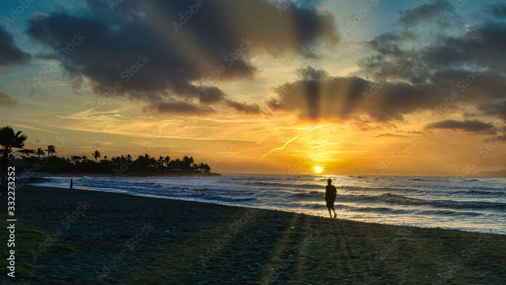 Fototapeta premium Silhouette of a man walking on a beach at sunrise with suns rays across the sky