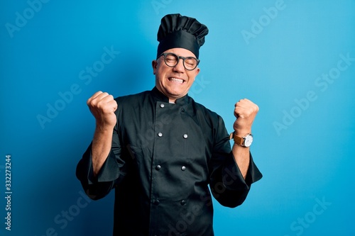 Middle age handsome grey-haired chef man wearing cooker uniform and hat very happy and excited doing winner gesture with arms raised, smiling and screaming for success. Celebration concept.