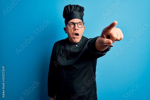 Middle age handsome grey-haired chef man wearing cooker uniform and hat Pointing with finger surprised ahead, open mouth amazed expression, something on the front