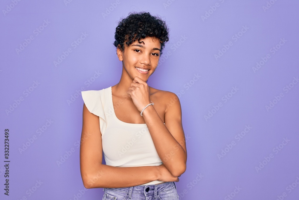 Young beautiful african american afro woman wearing casual t-shirt over purple background looking confident at the camera smiling with crossed arms and hand raised on chin. Thinking positive. Young beautiful african american afro woman wearing casual t-shirt over purple background looking confident at the camera smiling with crossed arms and hand raised on chin. Thinking positive.