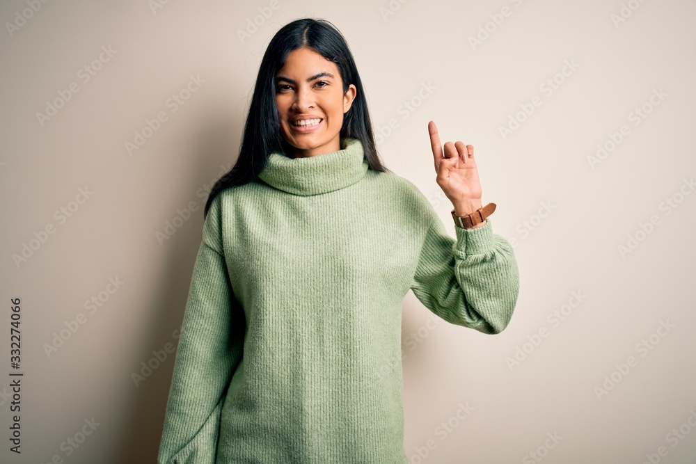 Young beautiful hispanic woman wearing green winter sweater over isolated background showing and pointing up with finger number one while smiling confident and happy. Young beautiful hispanic woman wearing green winter sweater over isolated background showing and pointing up with finger number one while smiling confident and happy.