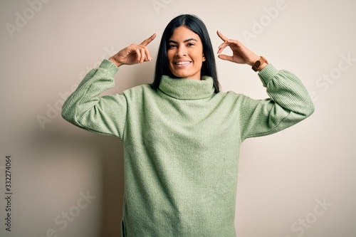 Young beautiful hispanic woman wearing green winter sweater over isolated background smiling pointing to head with both hands finger, great idea or thought, good memory © Krakenimages.com