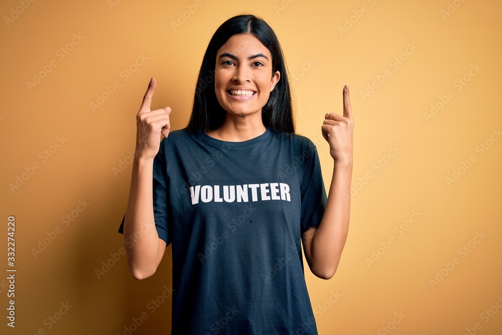 Young beautiful hispanic woman wearing volunteer t-shirt as social charity moral smiling amazed and surprised and pointing up with fingers and raised arms. Young beautiful hispanic woman wearing volunteer t-shirt as social charity moral smiling amazed and surprised and pointing up with fingers and raised arms.
