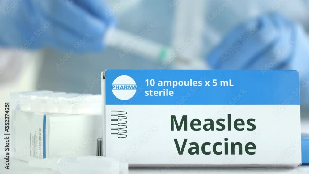 Ampoules with measles vaccine on the table near working laboratory assistant, fictional logo on the box Ampoules with measles vaccine on the table near working laboratory assistant, fictional logo on the box