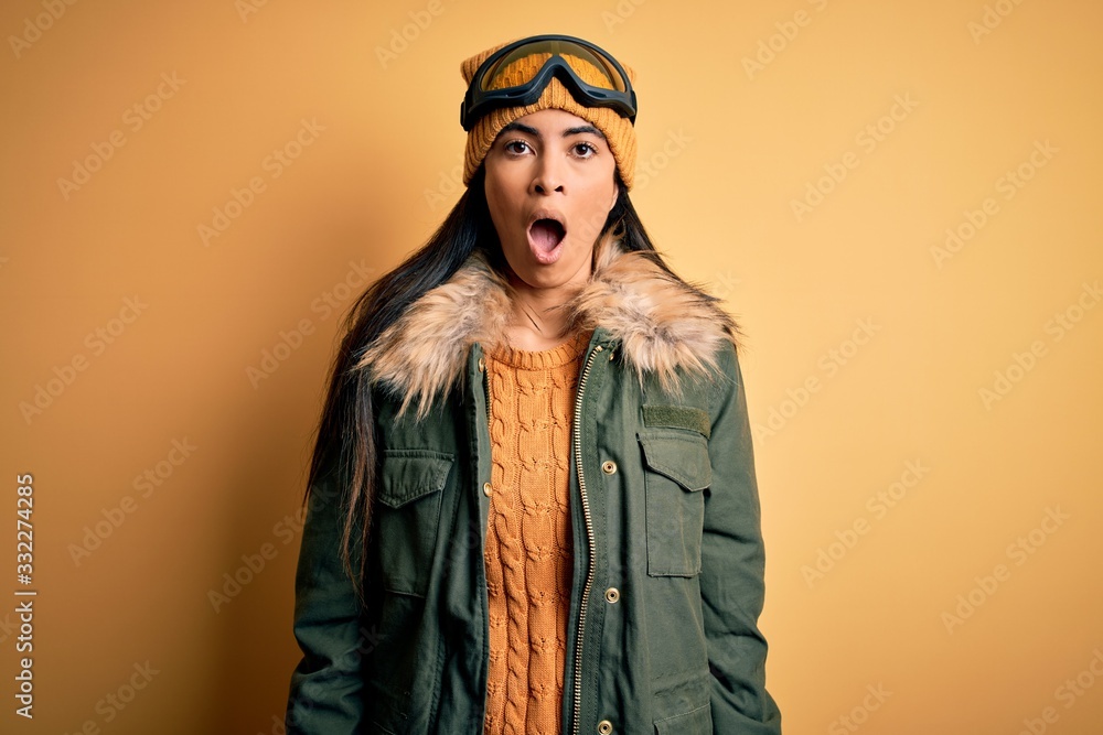 Young beautiful hispanic woman wearing ski glasses and coat for winter weather afraid and shocked with surprise and amazed expression, fear and excited face. Young beautiful hispanic woman wearing ski glasses and coat for winter weather afraid and shocked with surprise and amazed expression, fear and excited face.