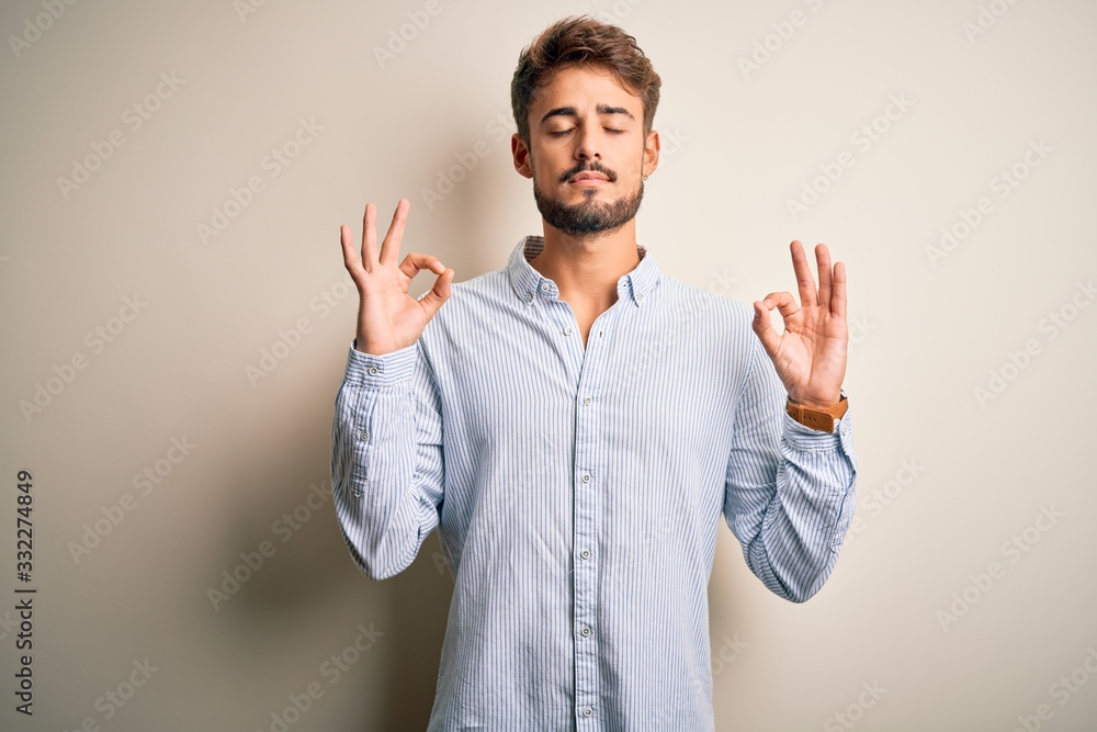 Young handsome man with beard wearing striped shirt standing over white background relaxed and smiling with eyes closed doing meditation gesture with fingers. Yoga concept. Young handsome man with beard wearing striped shirt standing over white background relaxed and smiling with eyes closed doing meditation gesture with fingers. Yoga concept.