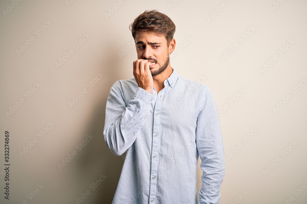 Young handsome man with beard wearing striped shirt standing over white background looking stressed and nervous with hands on mouth biting nails. Anxiety problem. Young handsome man with beard wearing striped shirt standing over white background looking stressed and nervous with hands on mouth biting nails. Anxiety problem.